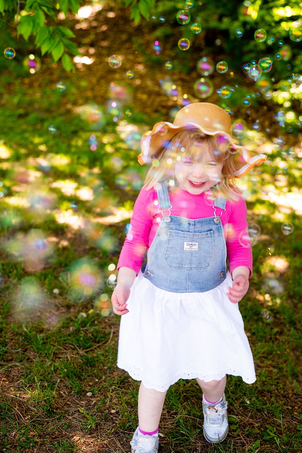 Soap bubbles float through a sunlit garden scene while a child in denim overalls and pink shirt plays below.
