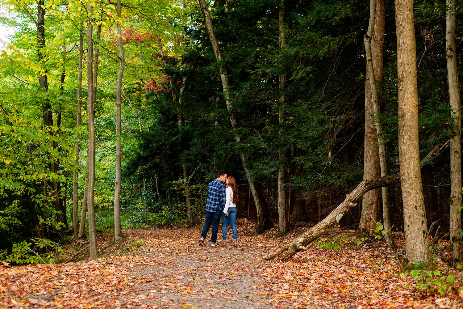 Couple strolls down leaf-covered forest path in autumn with tall trees and green foliage surrounding them.
