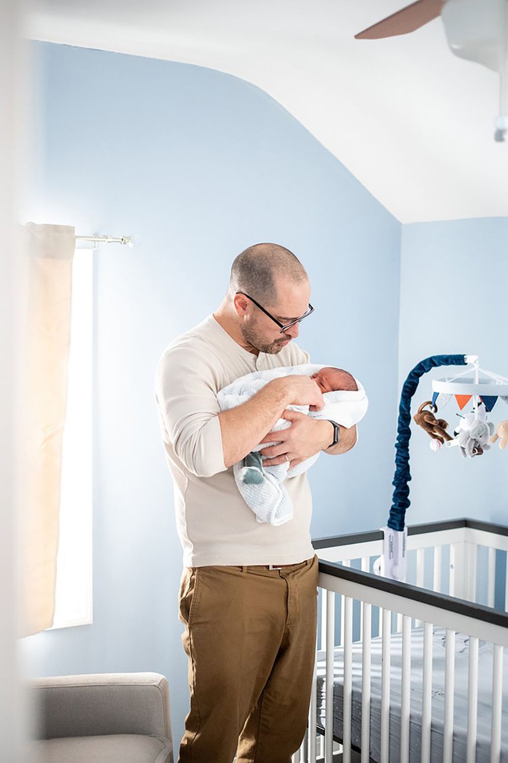 A person wearing glasses and khaki pants tenderly holds a newborn baby in a nursery with blue walls and a crib.