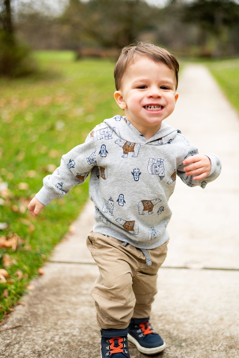 Small child wearing a bear-patterned hoodie and khakis runs joyfully on a sidewalk in a park setting.