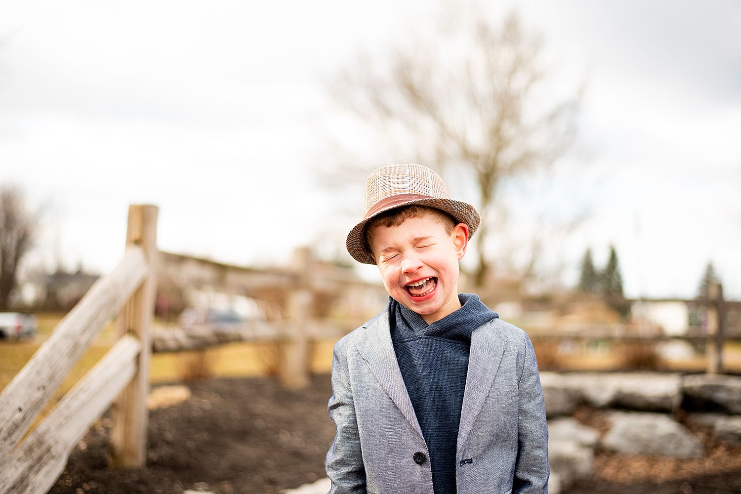 A young figure in a grey blazer and pink hat laughs joyfully outdoors on a cloudy day.