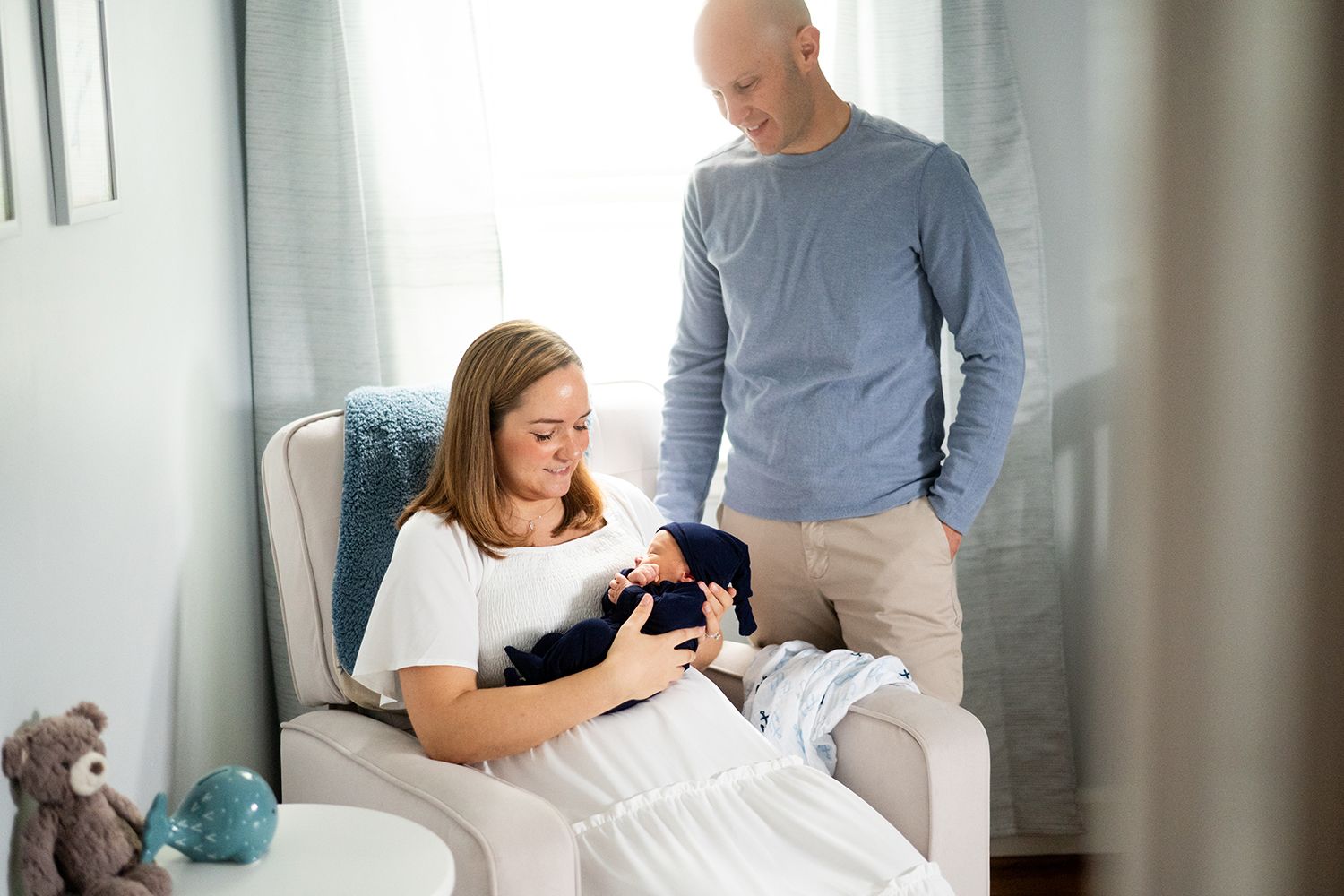 A couple sits together on a beige couch with newborn baby in bright living room with window light.