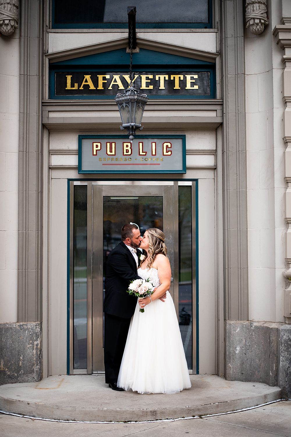A couple shares a romantic moment under the historic Lafayette Public entrance sign in an elegant stone doorway.