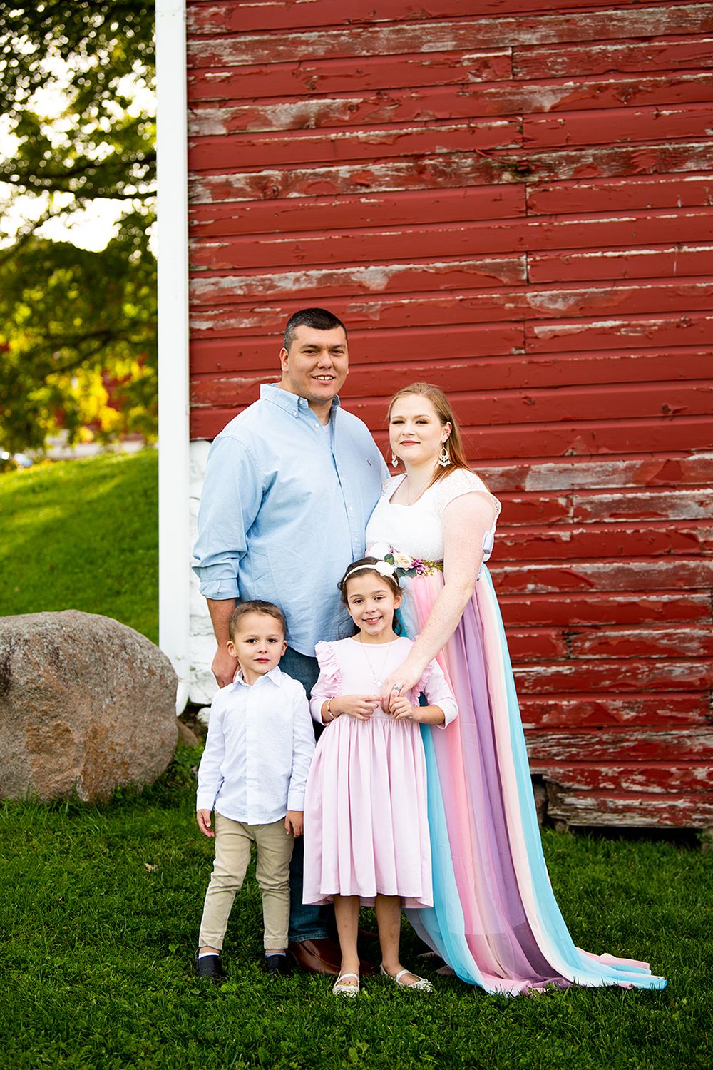 A family in coordinating blue and pink outfits poses together against a rustic red barn wall during an outdoor photo session.