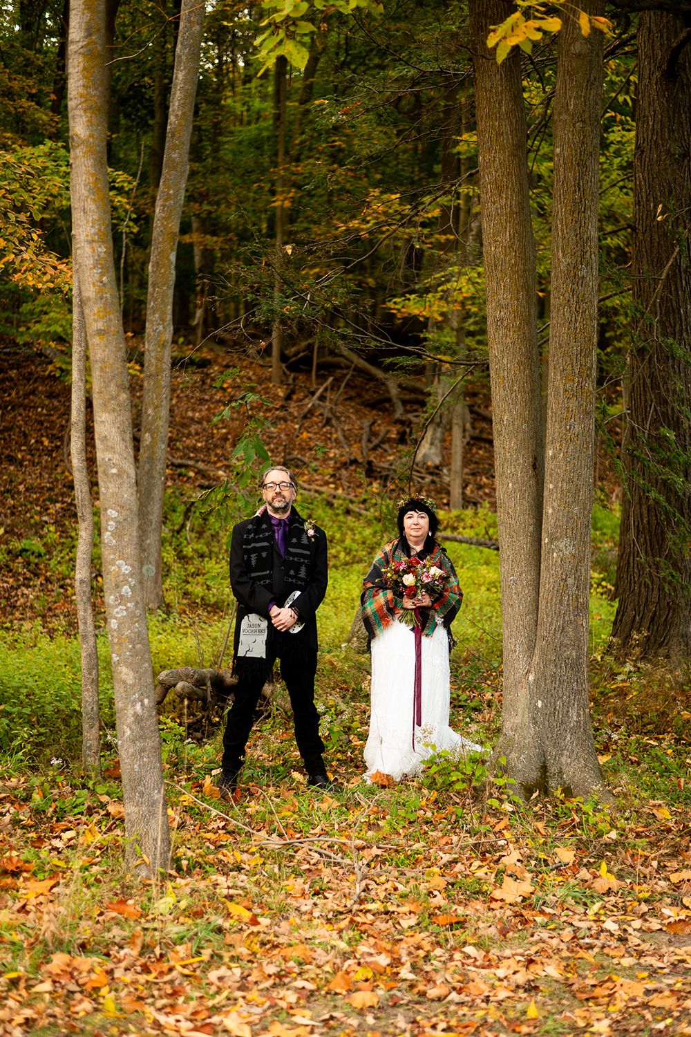 Two people stand among tall trees in an autumn forest with fallen leaves scattered on the ground.