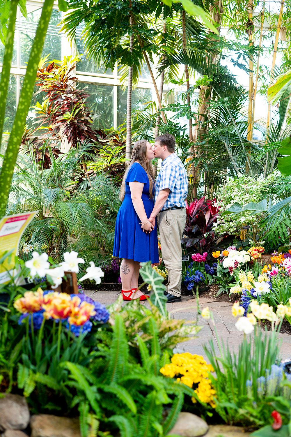 Two people share a romantic moment surrounded by vibrant tropical flowers and palm trees in a lush greenhouse garden.