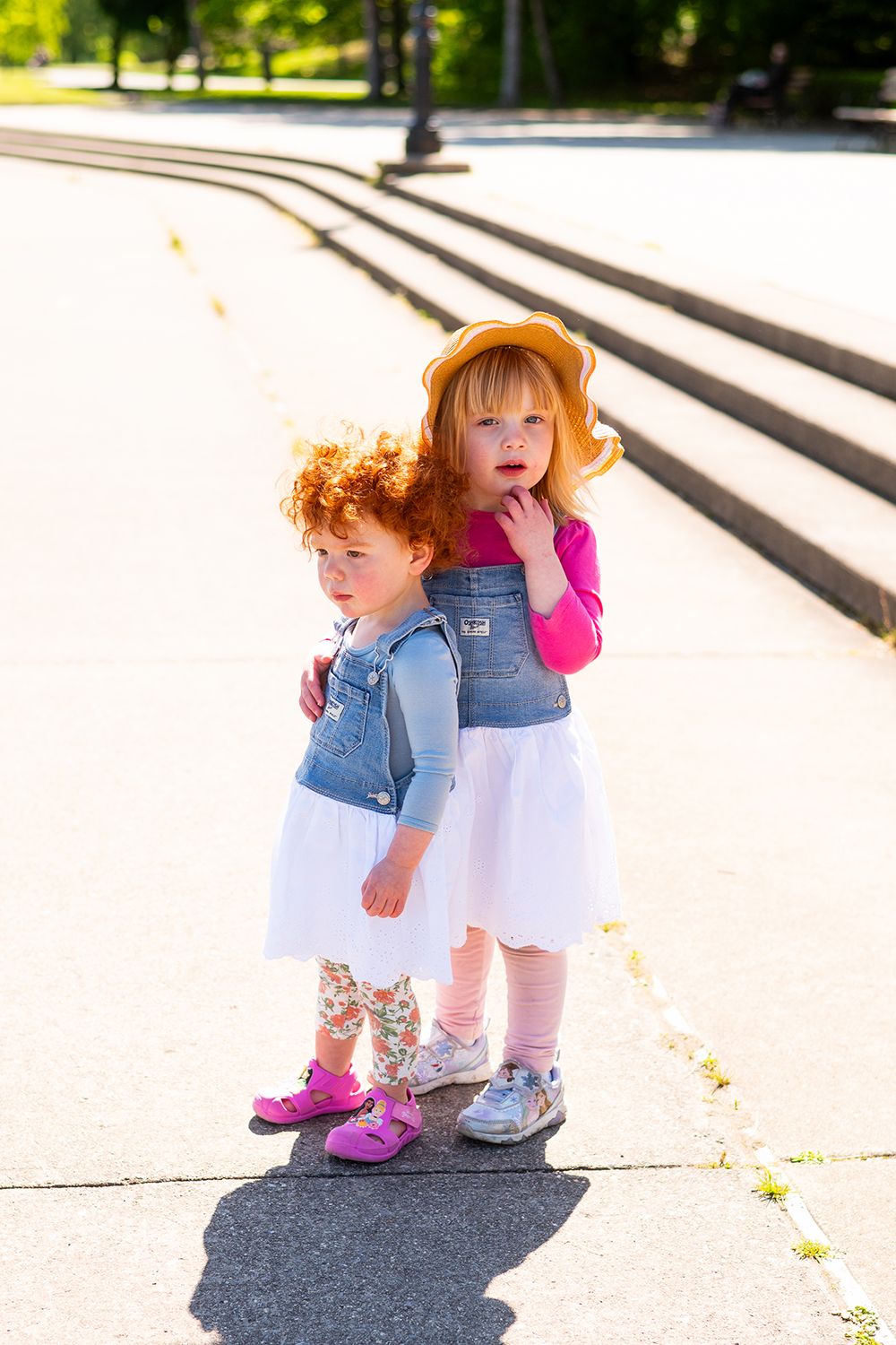 A small child in a denim jumper dress leans down to pick a dandelion on a sunny day.