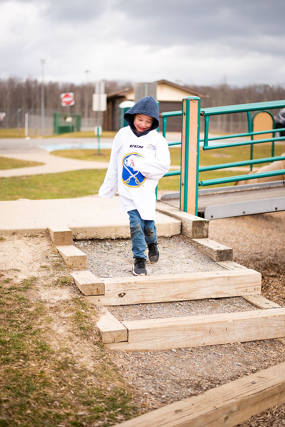 Child wearing a white sweatshirt and jeans descends wooden steps at an outdoor playground area on a cloudy day.