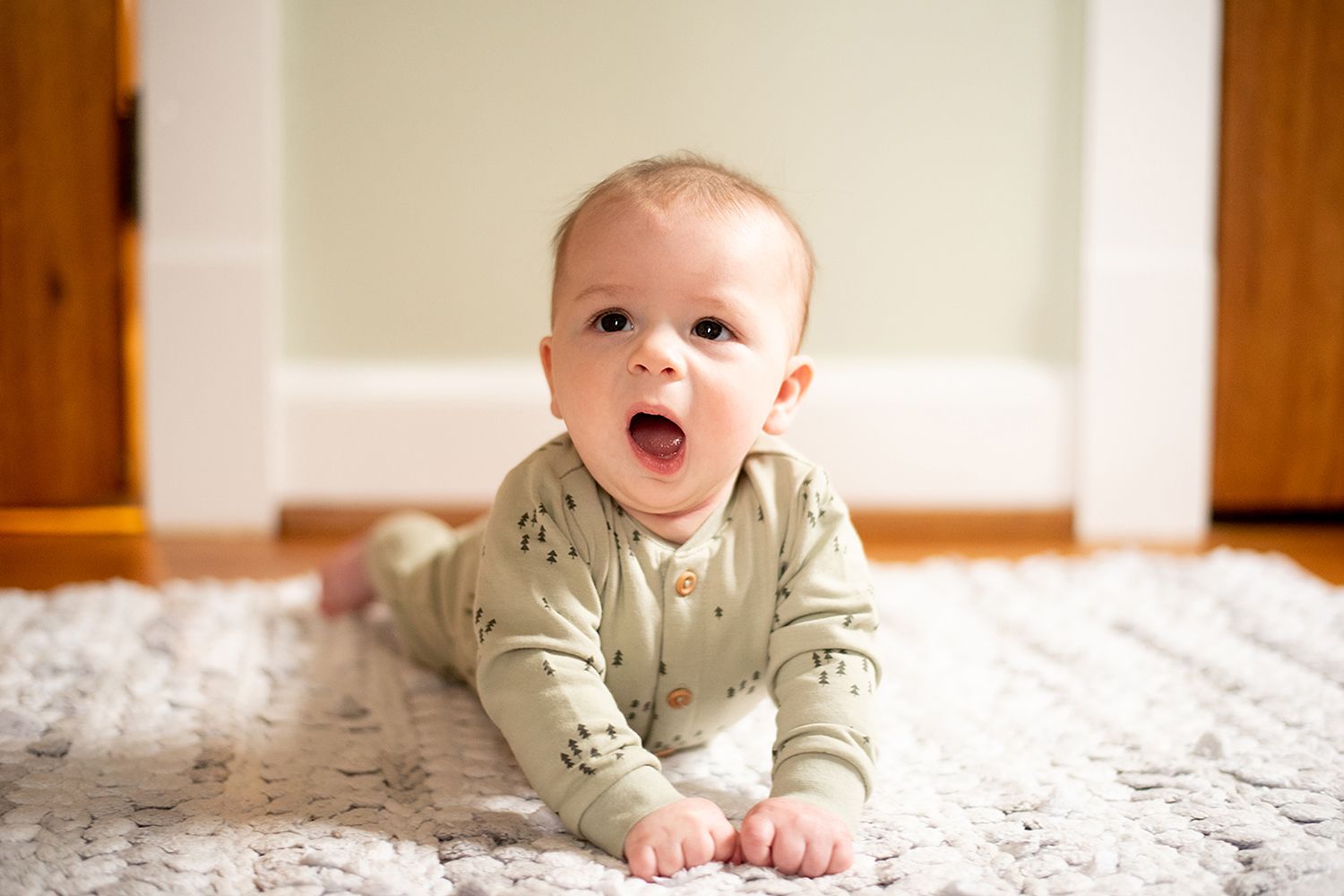 Baby in beige onesie lies on white rug with excited expression while learning to crawl.