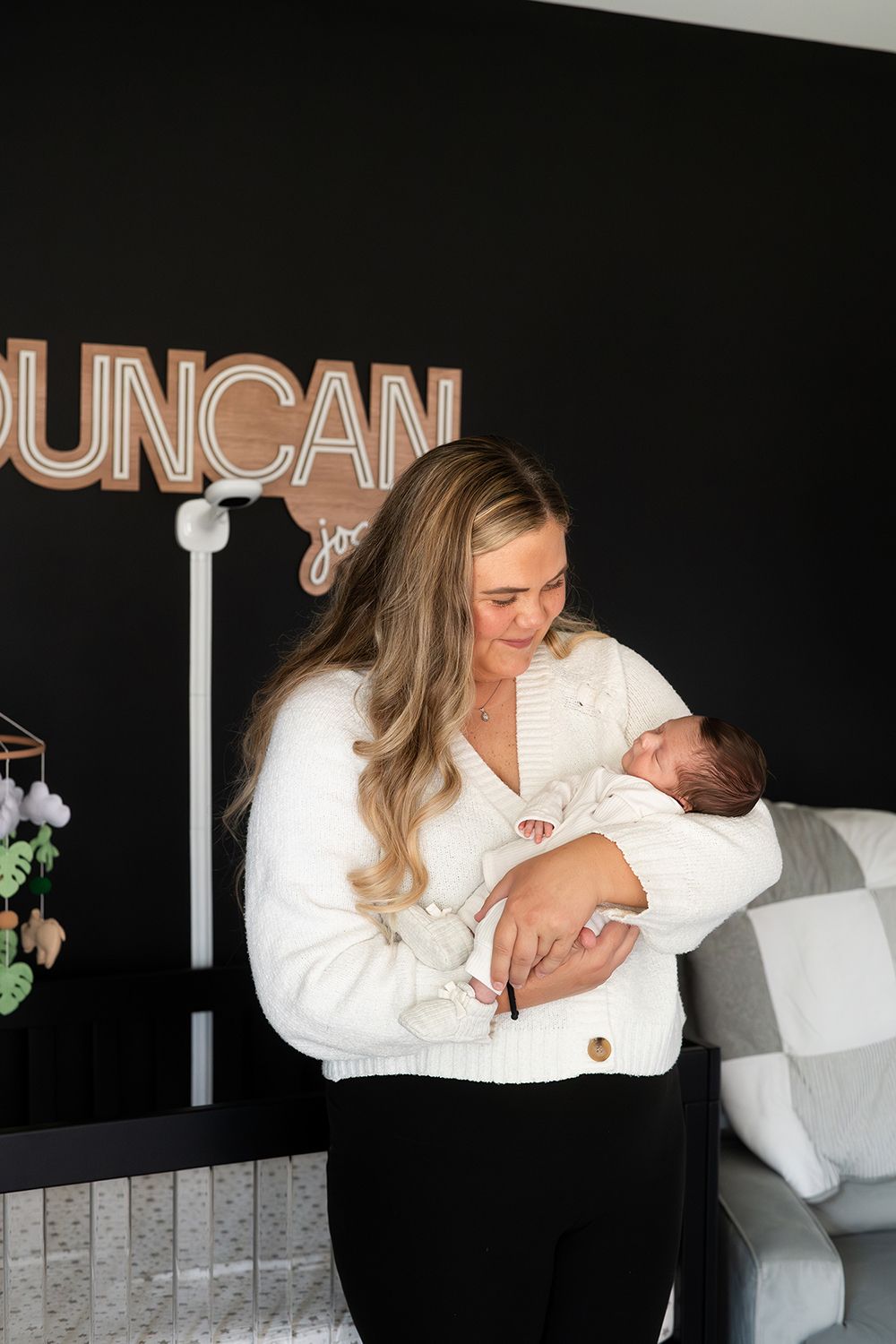 A person in white cardigan holding a newborn baby stands in front of a black wall with illuminated letters spelling Duncan.