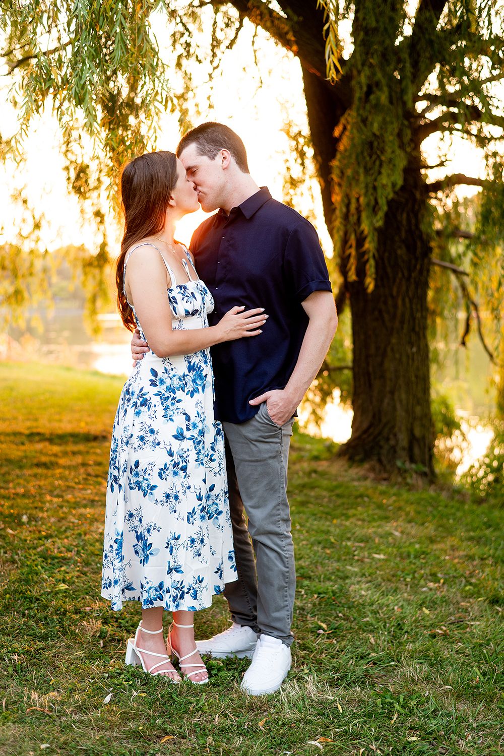 A couple shares a tender moment in a park at sunset with willow trees creating a romantic backdrop.