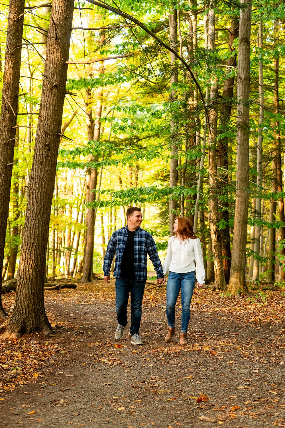 A couple walks together on an autumn path through a sunlit forest with tall trees and fallen leaves.