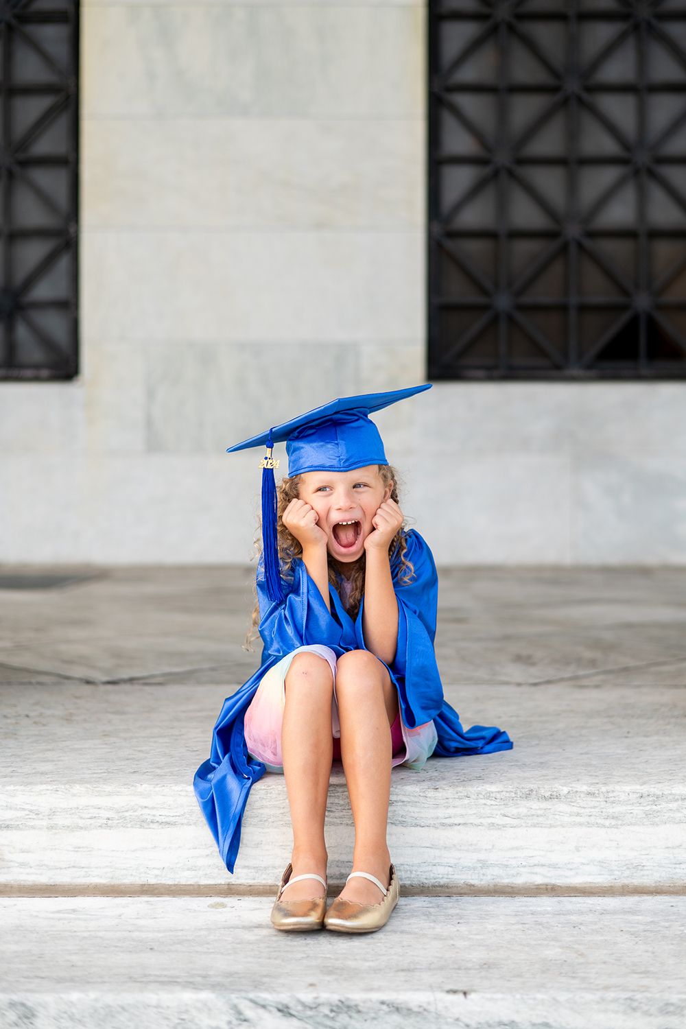 Graduate in blue cap and gown sits on stone steps displaying pure joy and excitement.