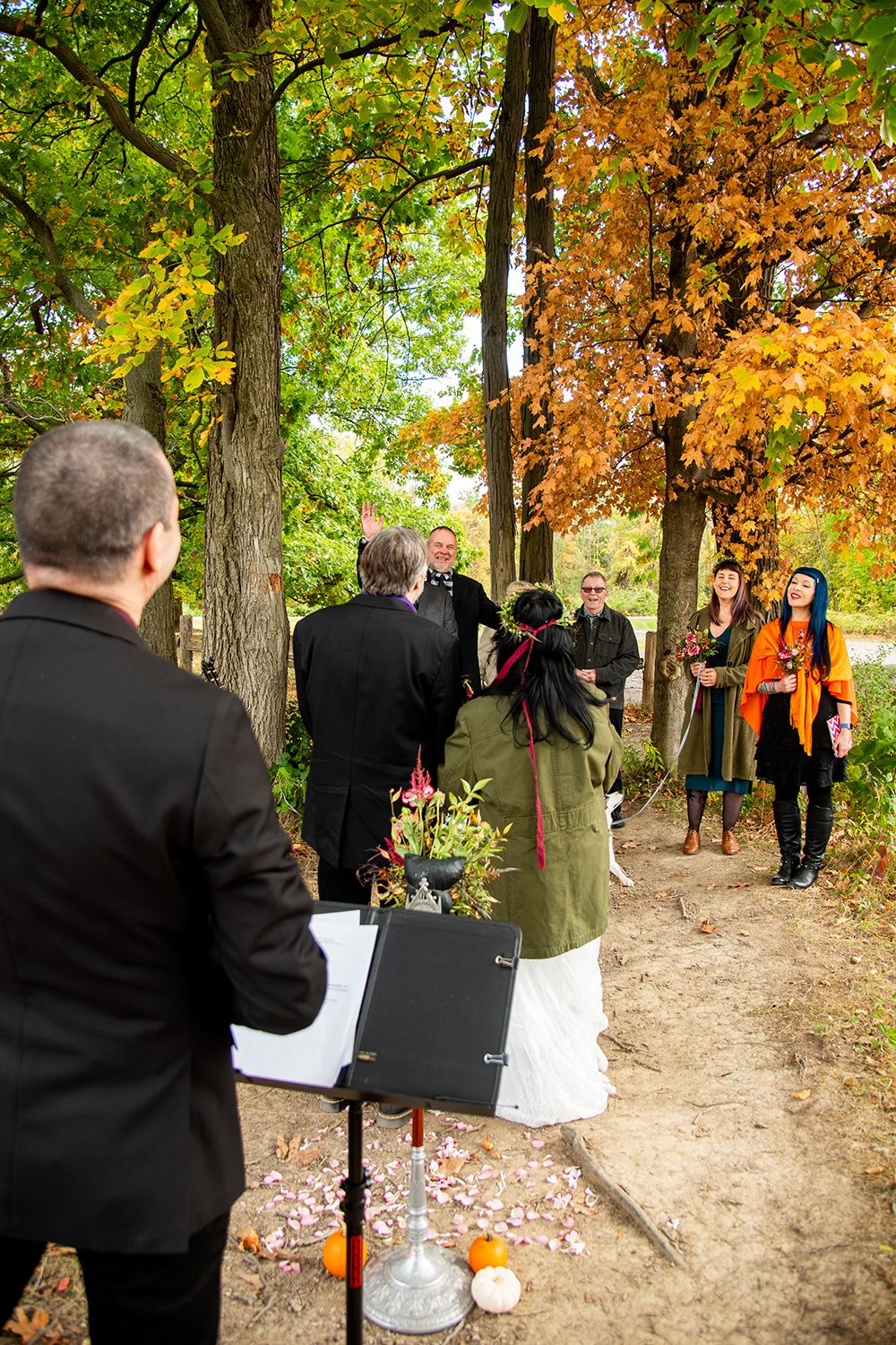Wedding guests gather on a wooded trail during an autumn ceremony with orange and yellow leaves overhead.