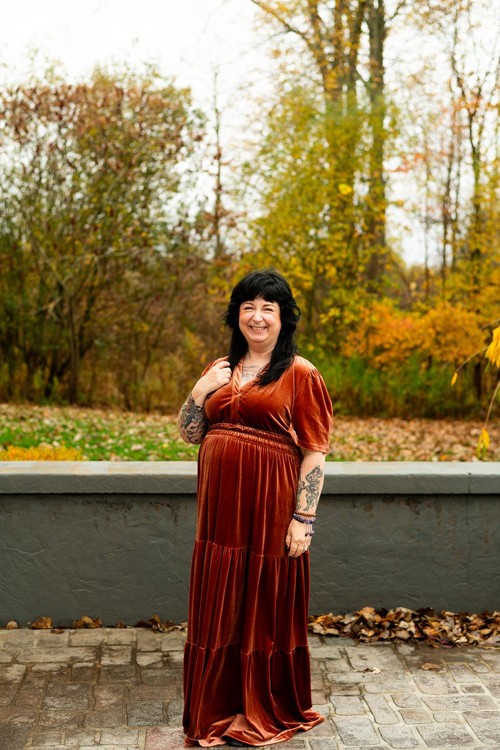 Person in burnt orange velvet maxi dress poses on stone patio surrounded by autumn foliage.