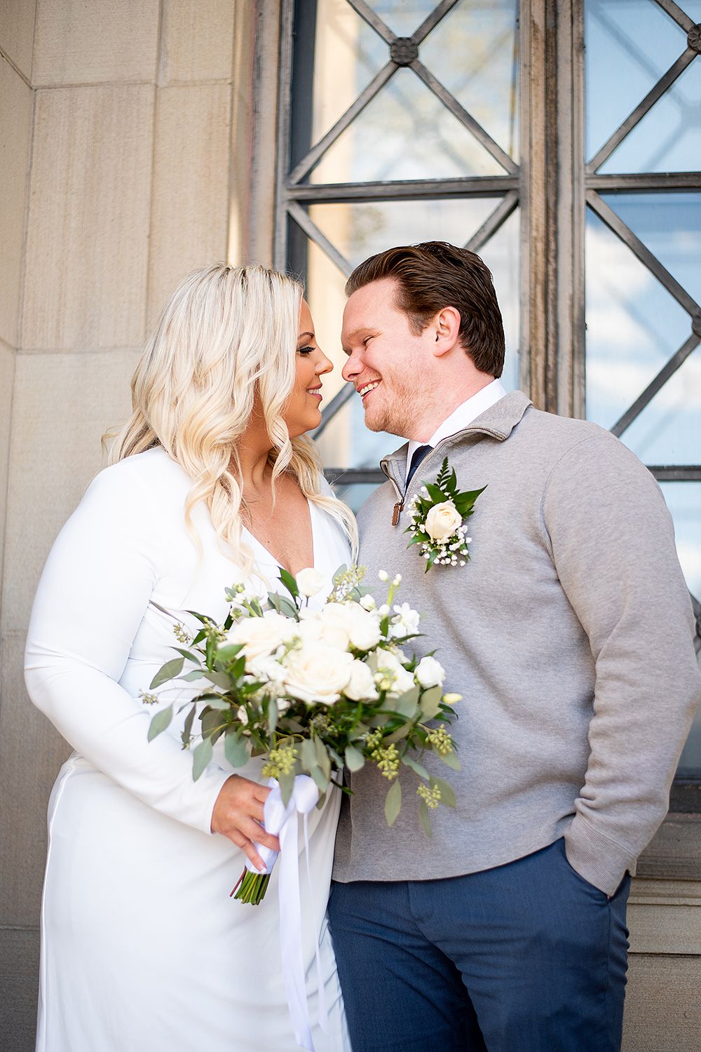Newlywed couple shares intimate moment next to paned window while bride holds white floral bouquet and greenery.