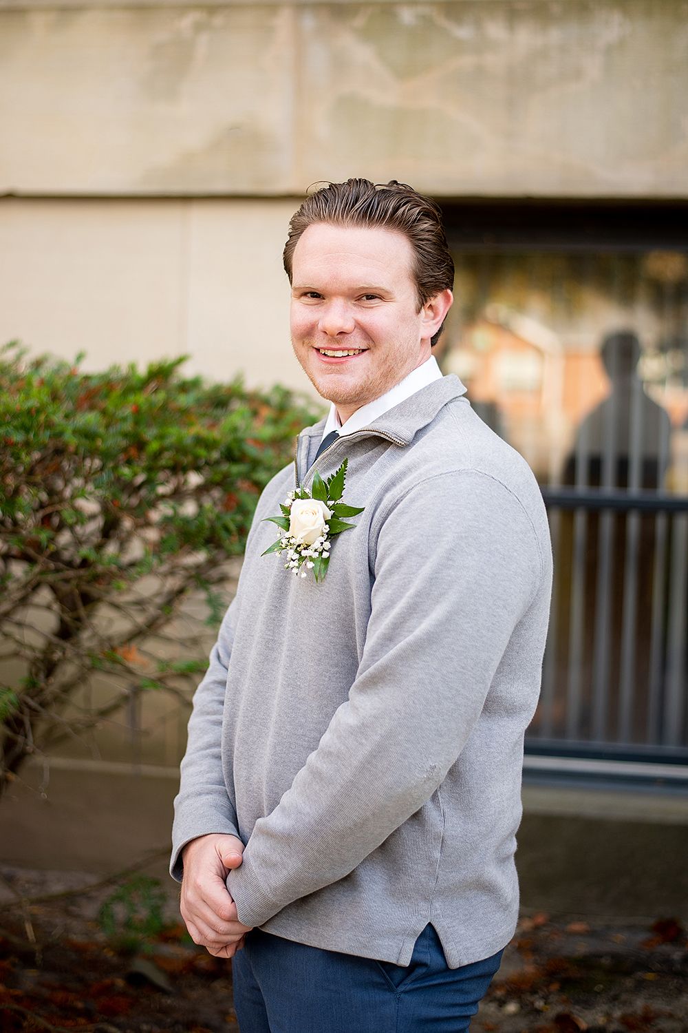A person wearing a gray sweater and dark pants posing with a white boutonniere against an outdoor background.