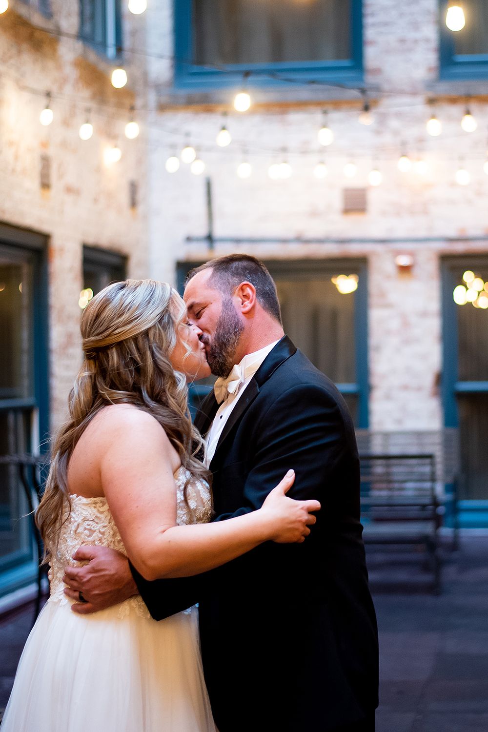 Three romantic photos of a couple sharing intimate kisses in a charming industrial courtyard setting with string lights.