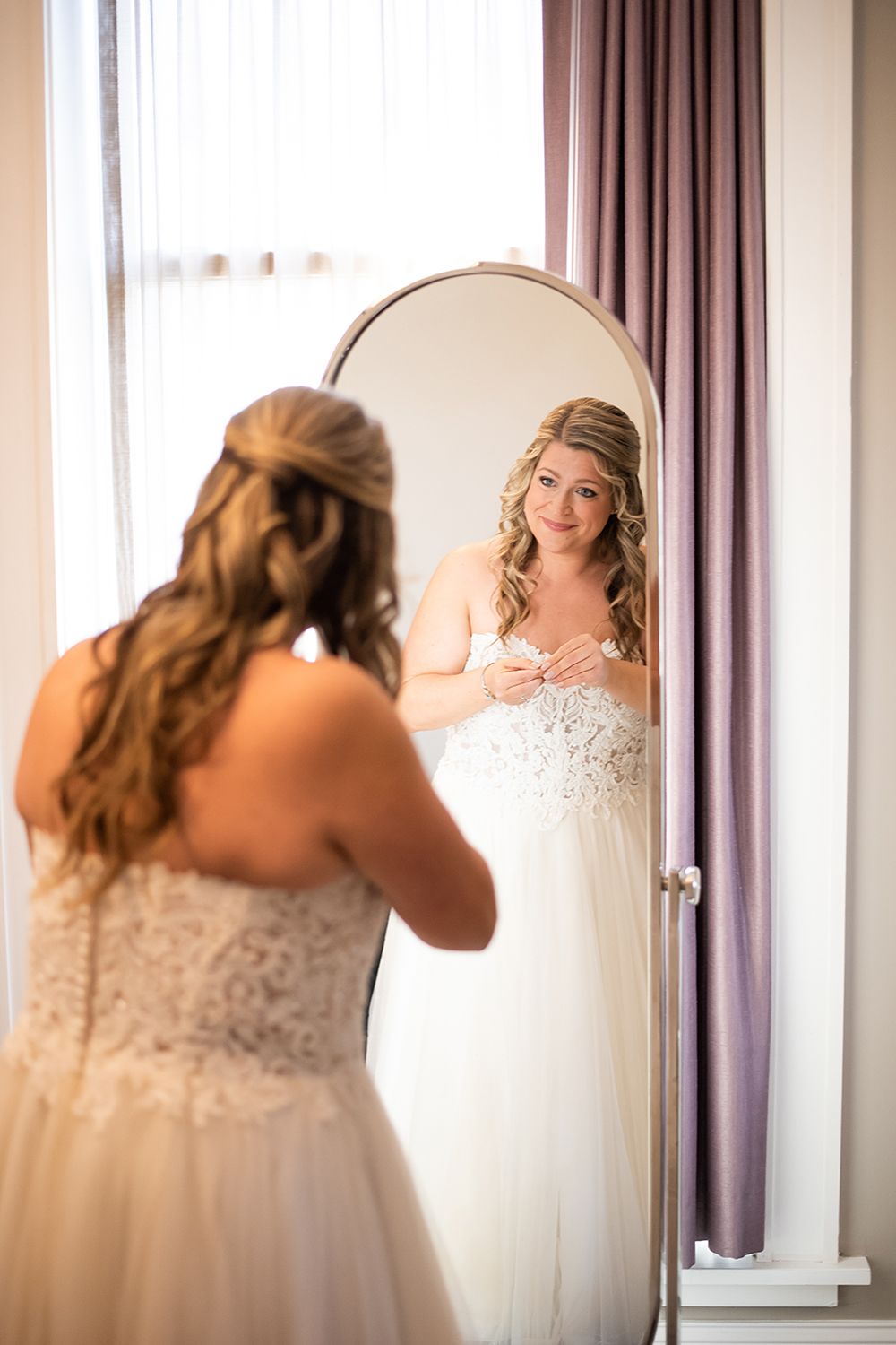 A white wedding dress is reflected in an oval mirror with purple curtains in the background.