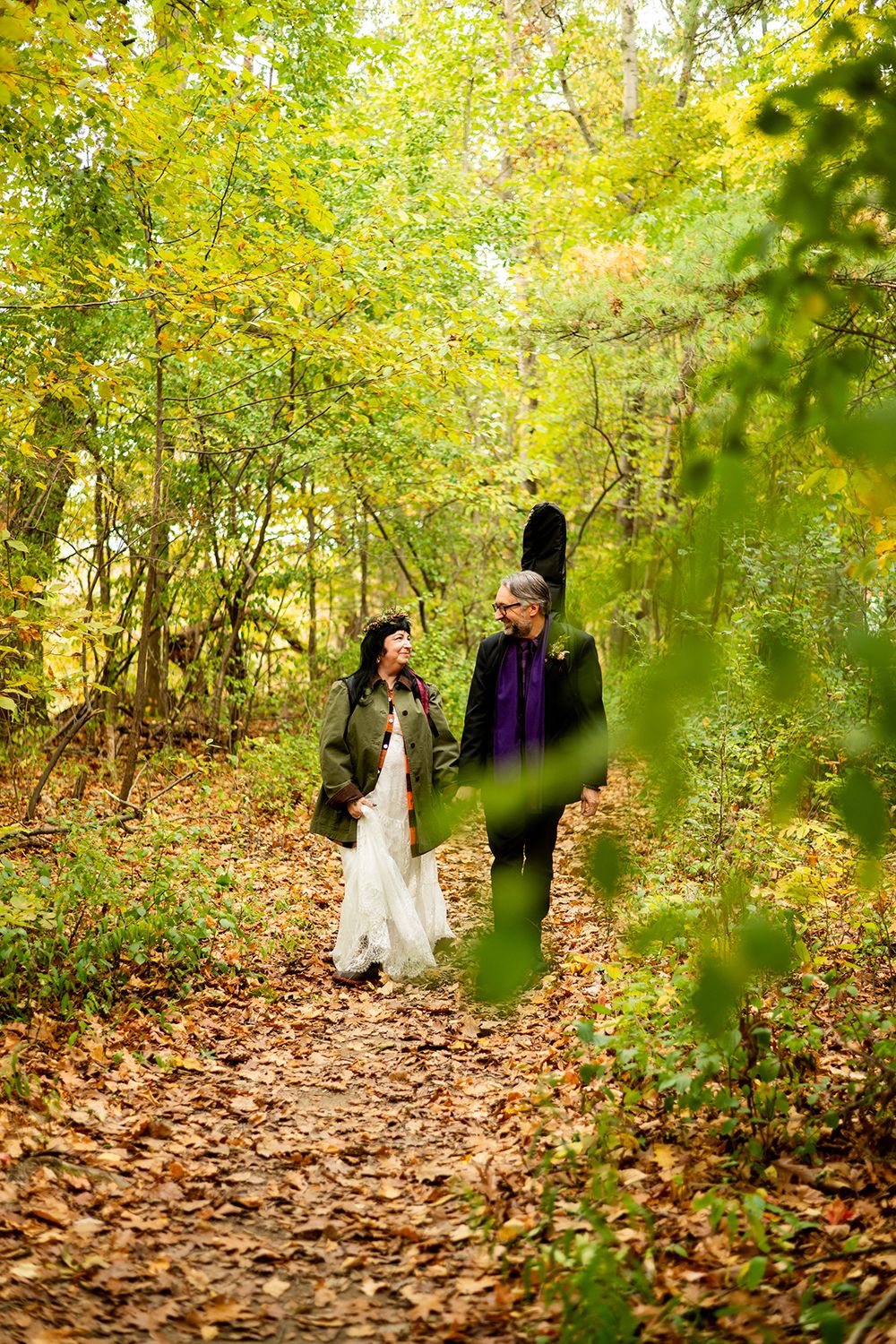 A couple walks together on a path covered in fall leaves surrounded by yellow and green autumn foliage.