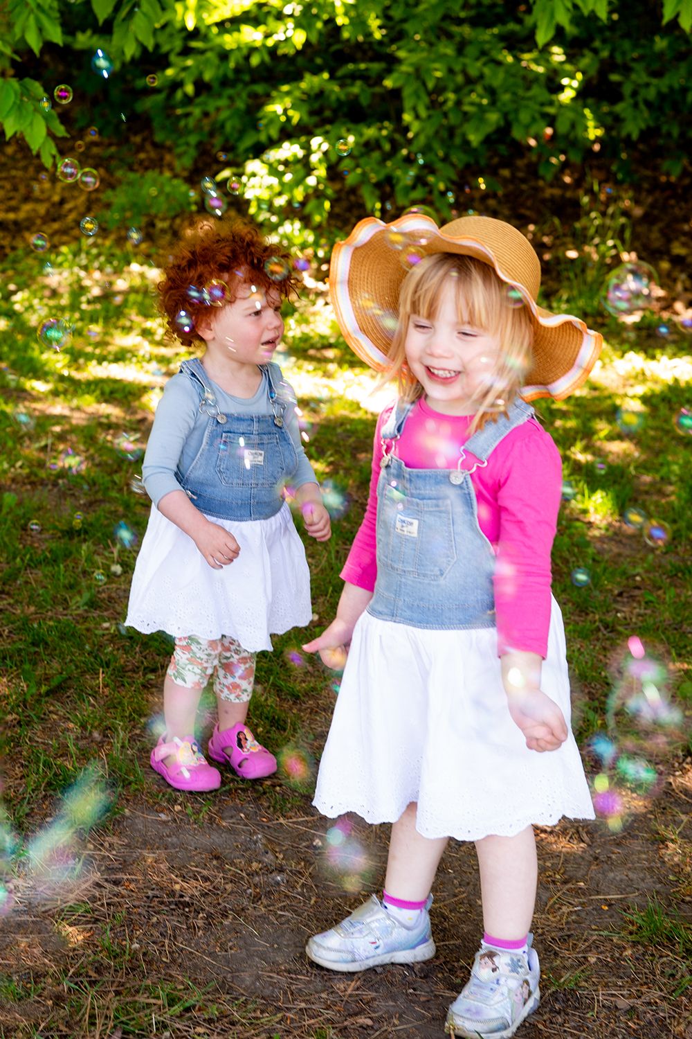 Two young children in matching jean dress overalls and white skirts play outdoors in a garden setting during summertime.