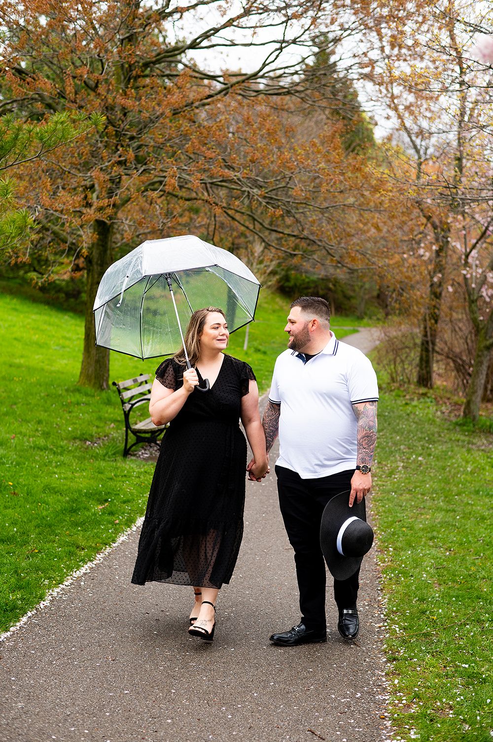 Couple walking together on a paved path through autumn trees, with one person holding a clear umbrella.