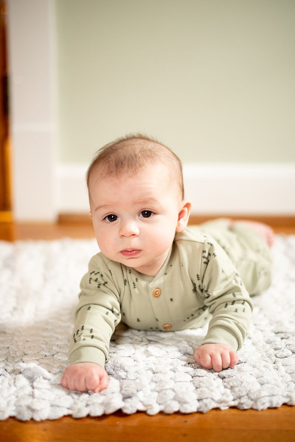 Baby in light green printed onesie lies on textured white rug during indoor photo session.