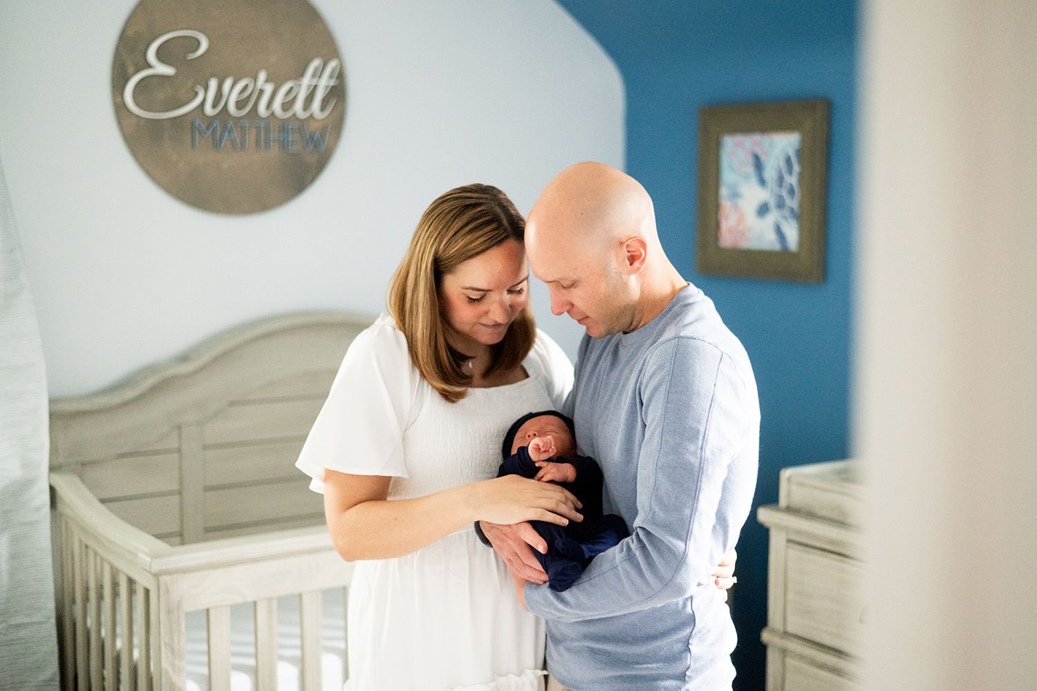 A couple stands together in a nursery with blue walls and a white crib, looking down lovingly at their newborn.