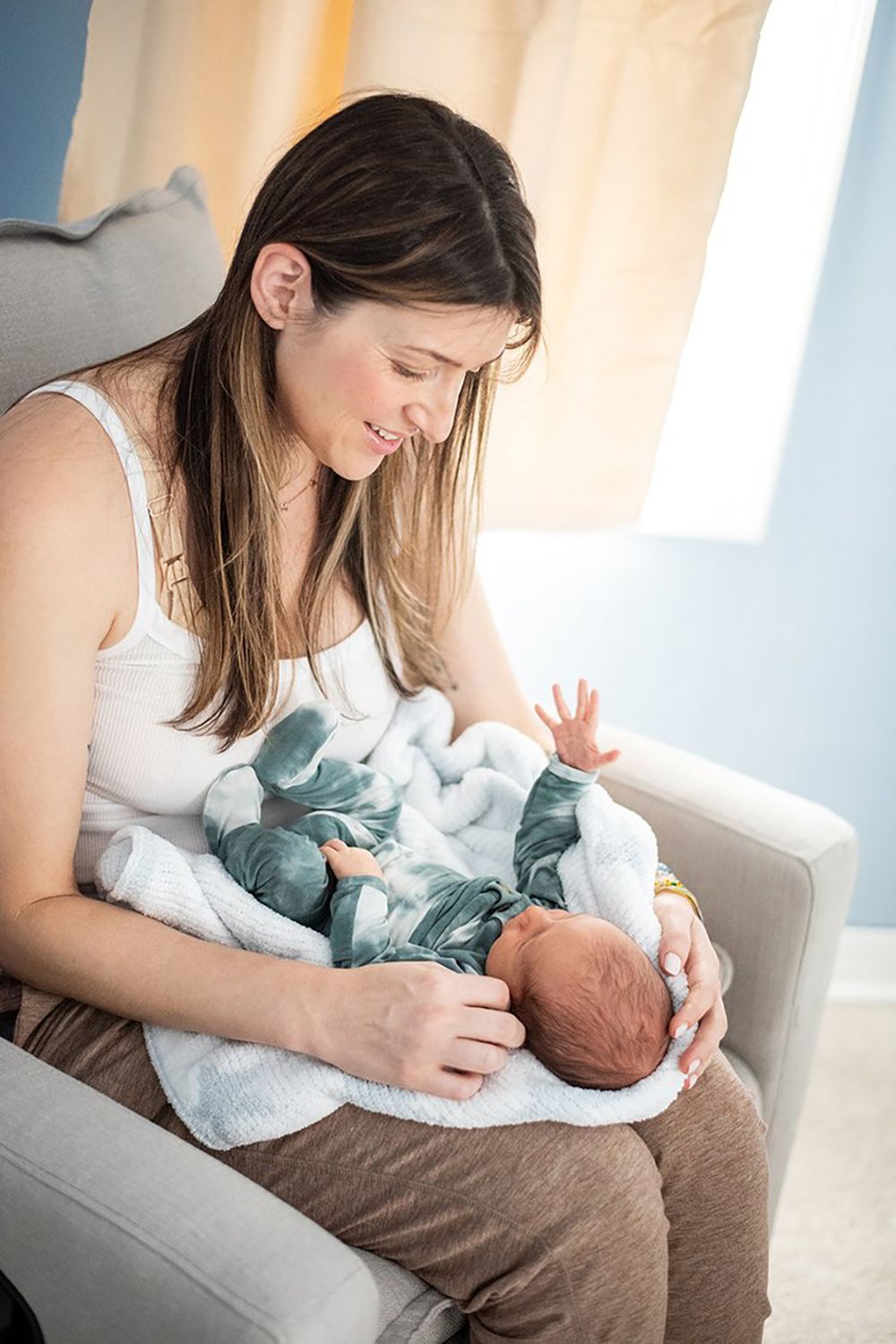 A caregiver in a white tank top holds a swaddled newborn, sitting in a chair near a sunlit window.