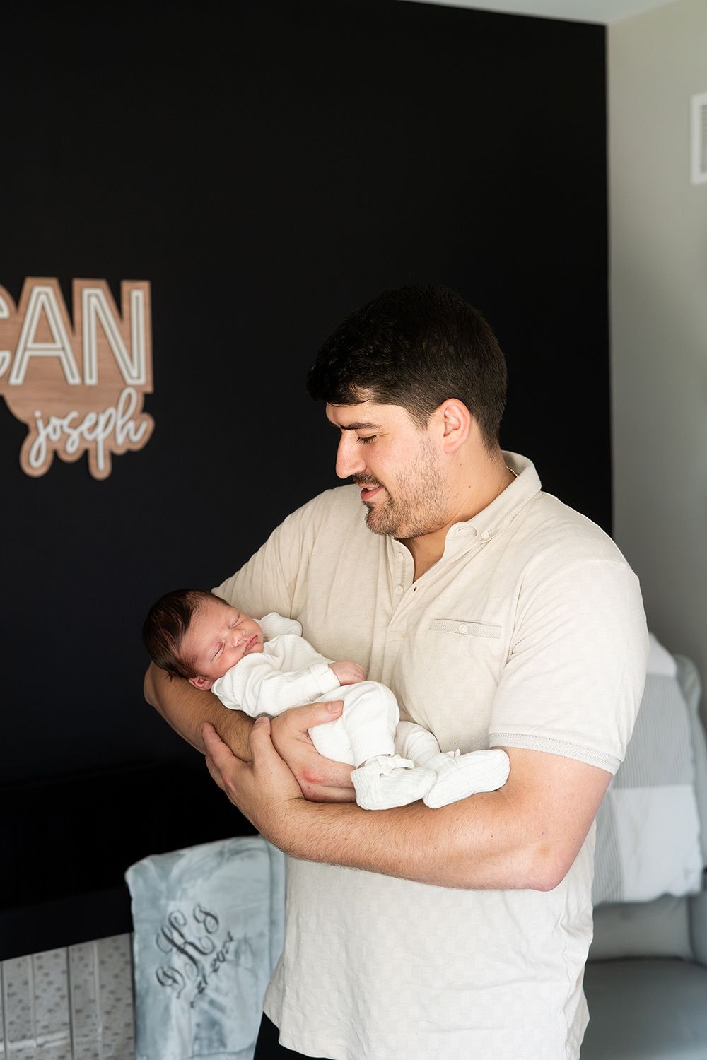 An adult in a beige shirt tenderly holds a newborn baby swaddled in white against a dark wall with text decor.