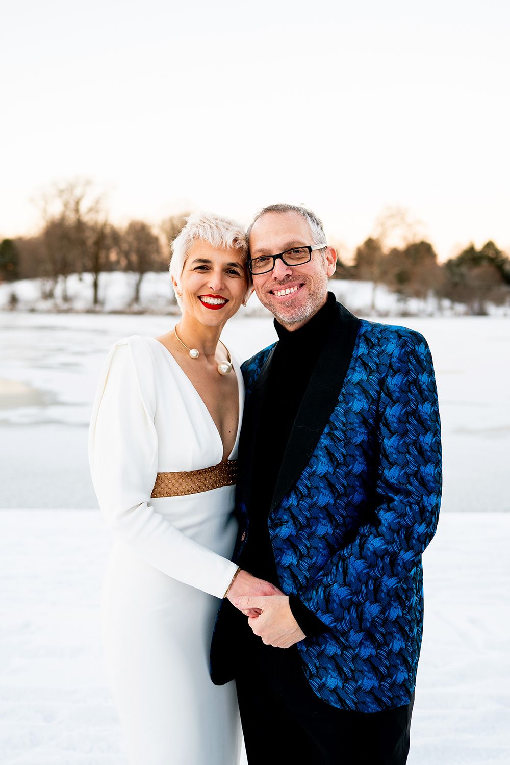 A couple embraces in a snowy winter landscape wearing contrasting white and blue outfits at sunset.
