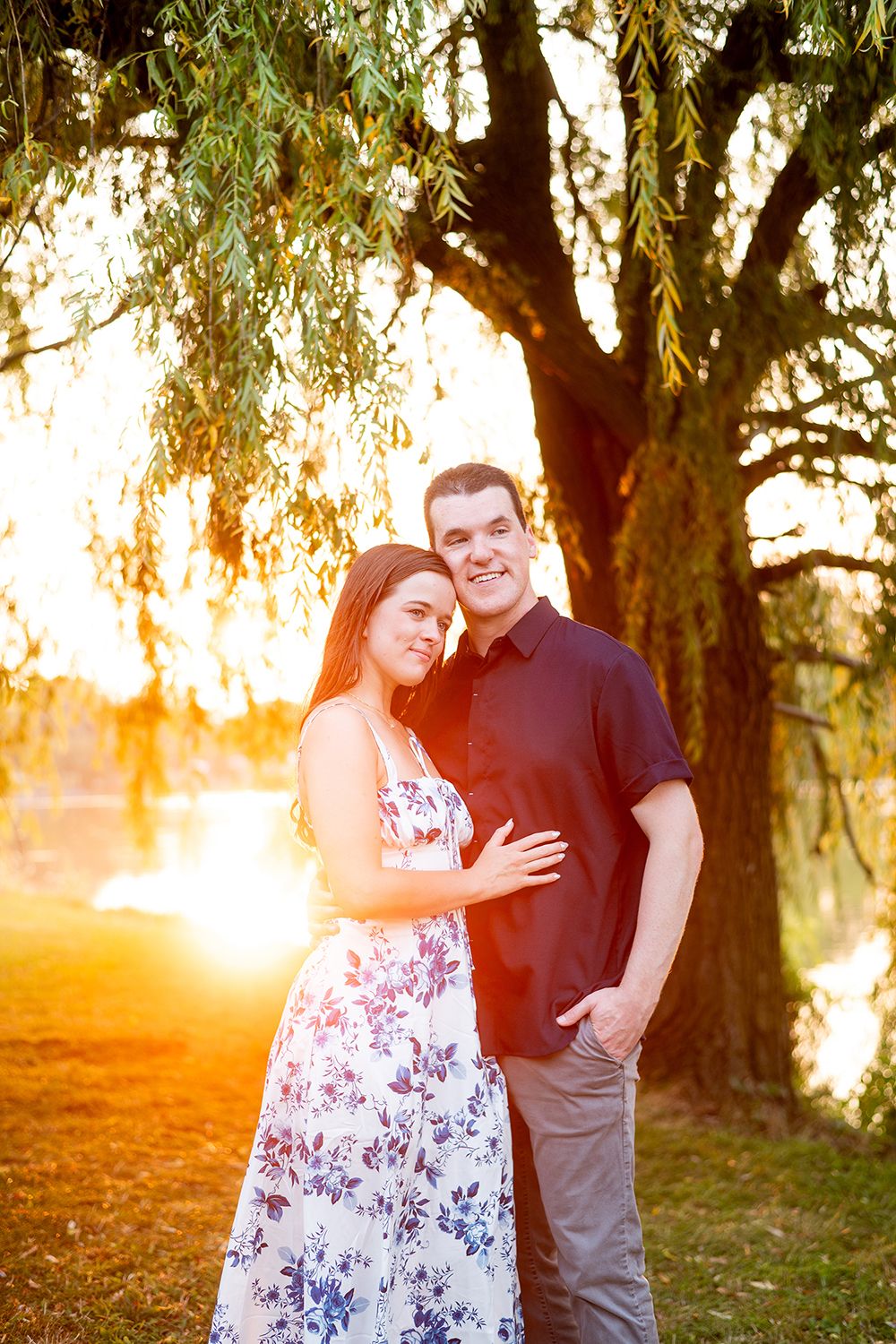 A couple embraces under a willow tree at sunset with warm golden light filtering through the branches.