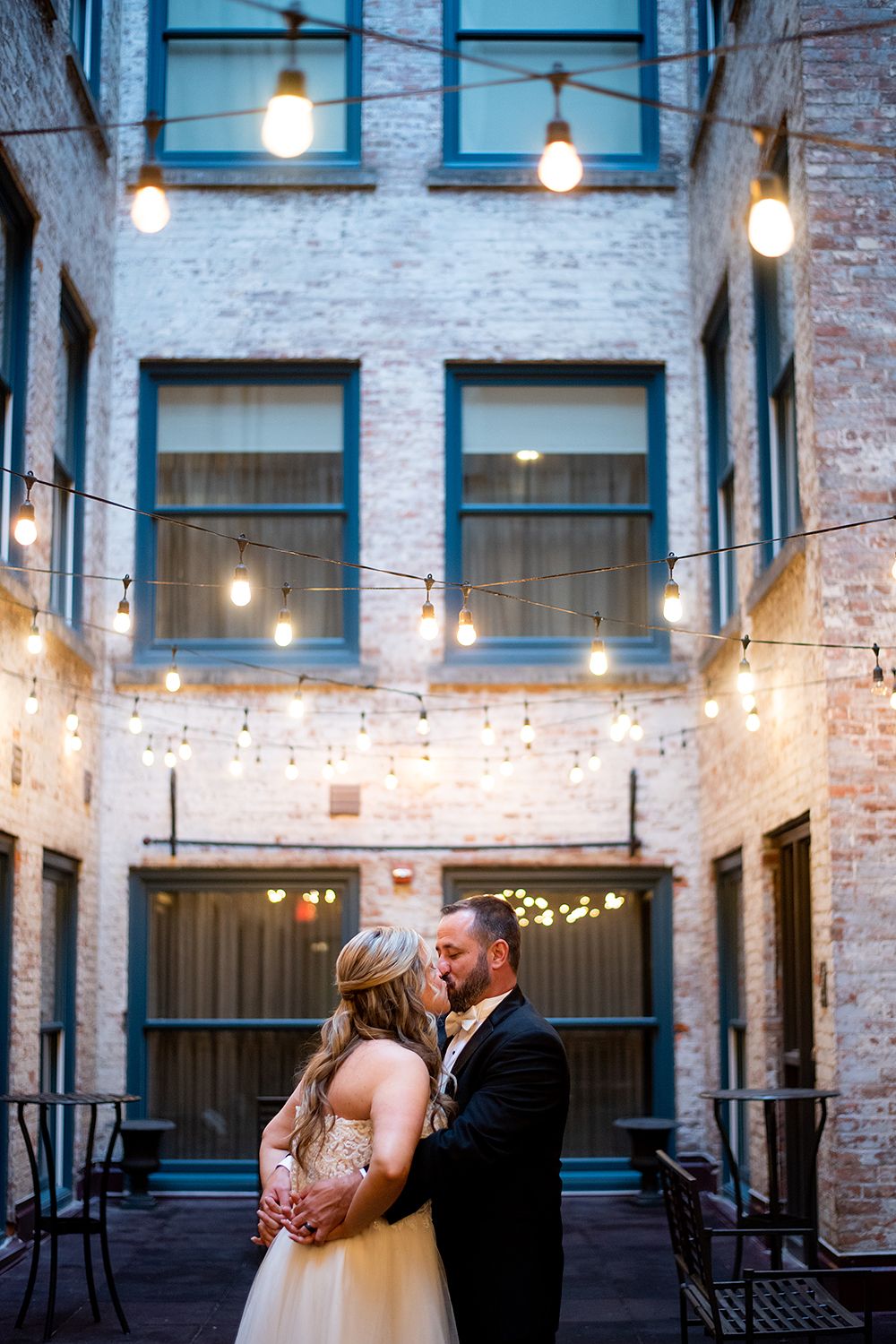 Couple shares romantic moment under string lights in an industrial brick courtyard with windows at dusk.