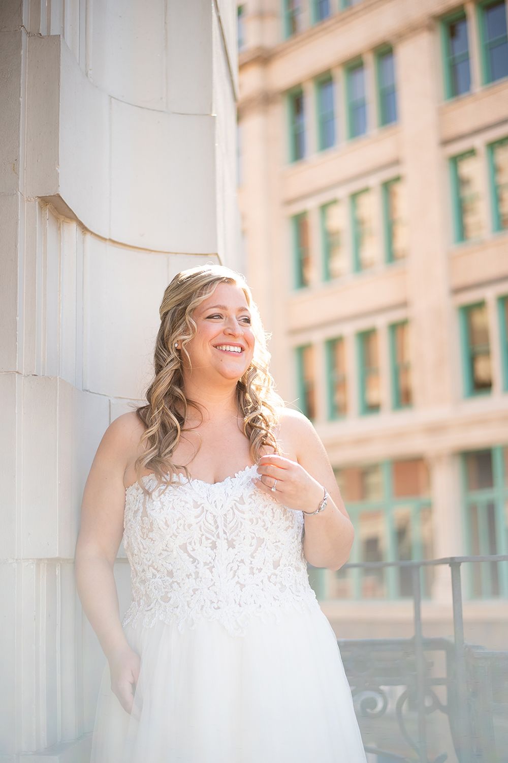 A bride in a white lace strapless wedding gown poses against a light stone column with a building in the background.