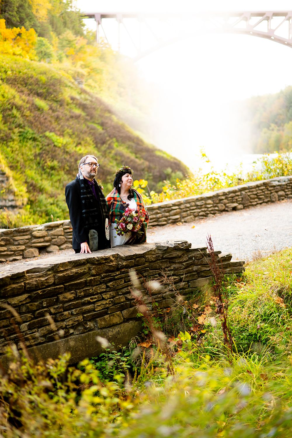 A couple sits on a rustic stone wall surrounded by autumn foliage as golden sunlight streams through the scene.