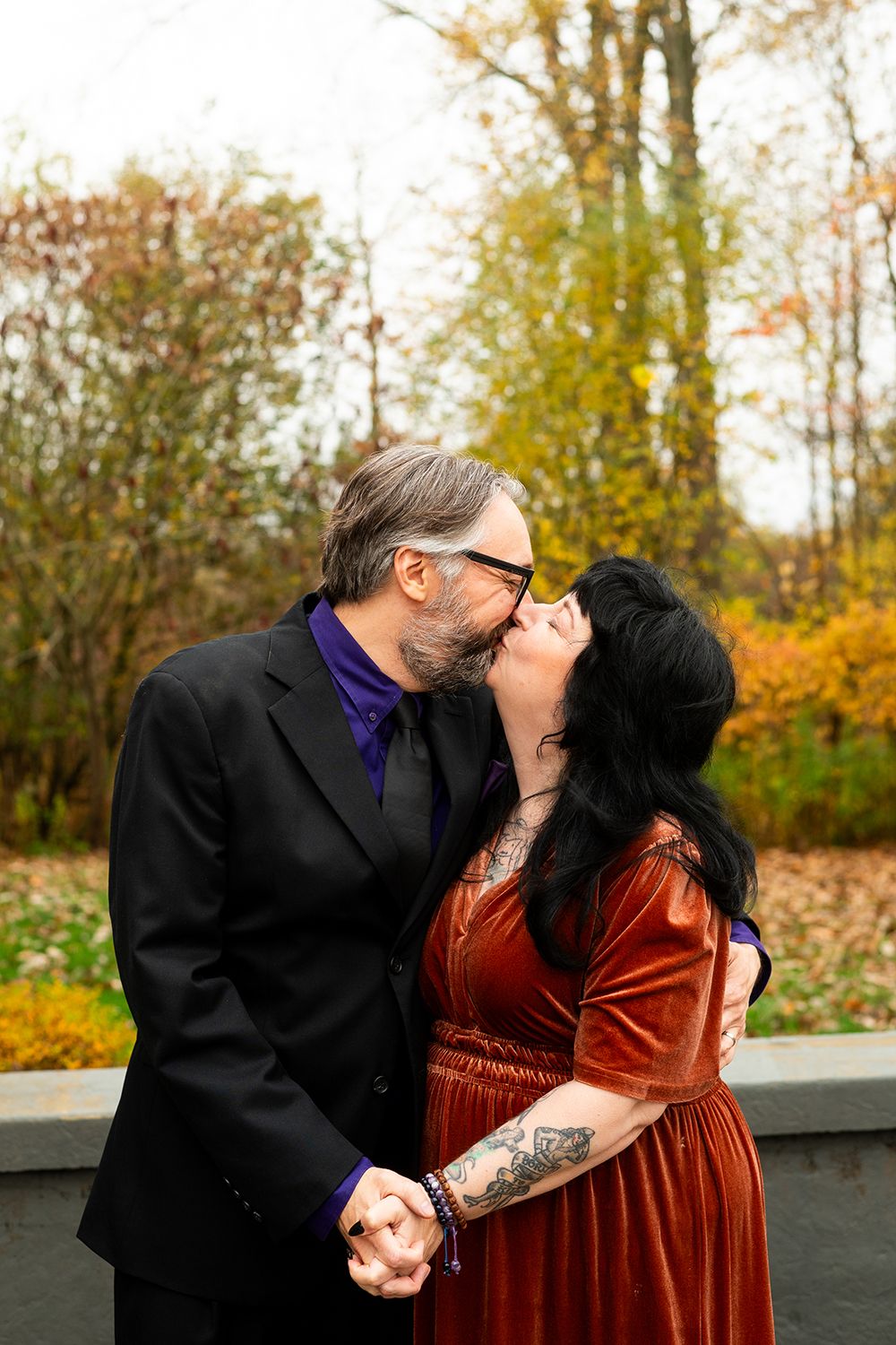 A couple shares tender moments in an outdoor autumn setting wearing black and burgundy outfits.