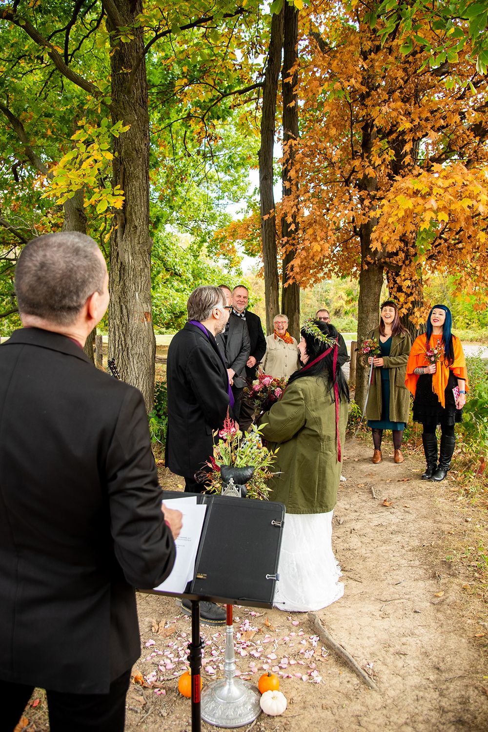 A group gathers for an outdoor autumn ceremony among trees with orange and green foliage.