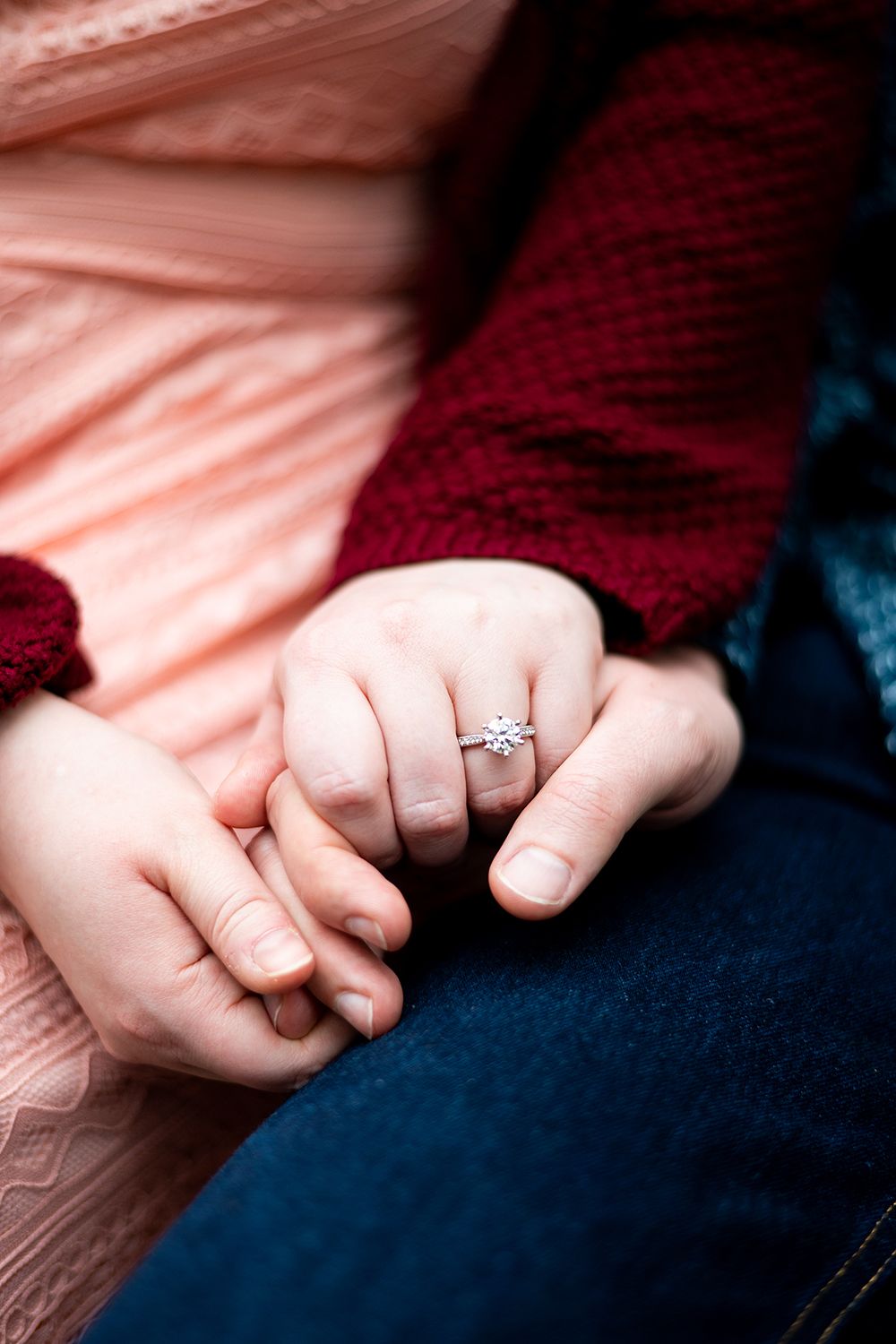Close-up of clasped hands with an engagement ring, against pink, red and blue textured fabric backdrop.