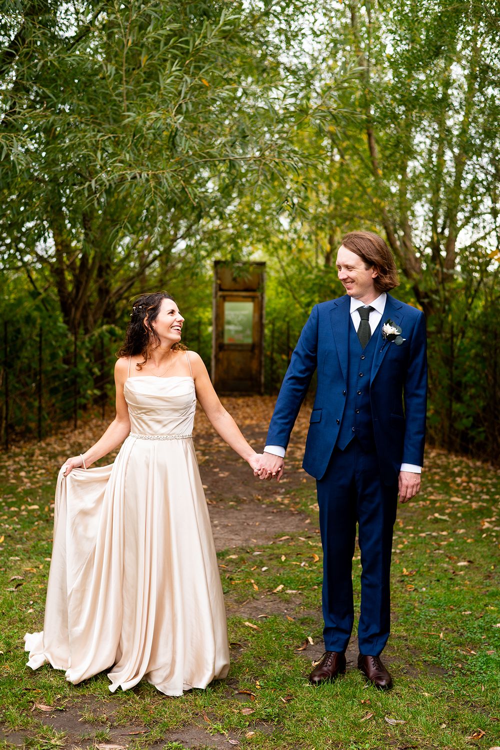 A couple in a wedding gown and navy suit holds hands in an outdoor garden setting with green foliage and autumn leaves.
