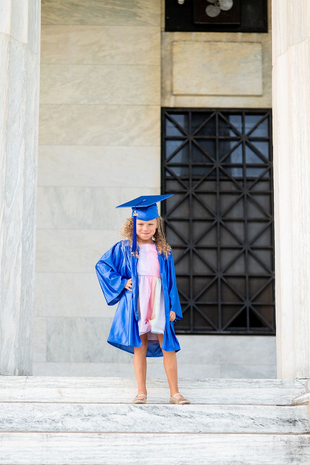 Graduate poses confidently in blue regalia and pink dress on marble steps of institutional building.
