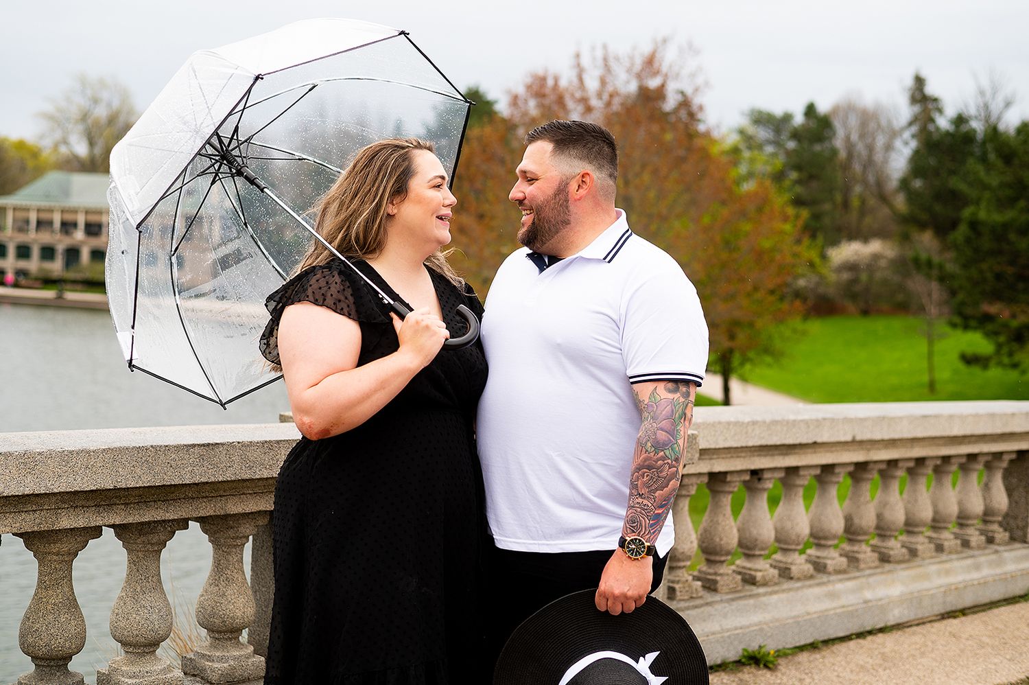 A couple share a joyful moment together on a stone balcony while holding a clear umbrella on an autumn day.
