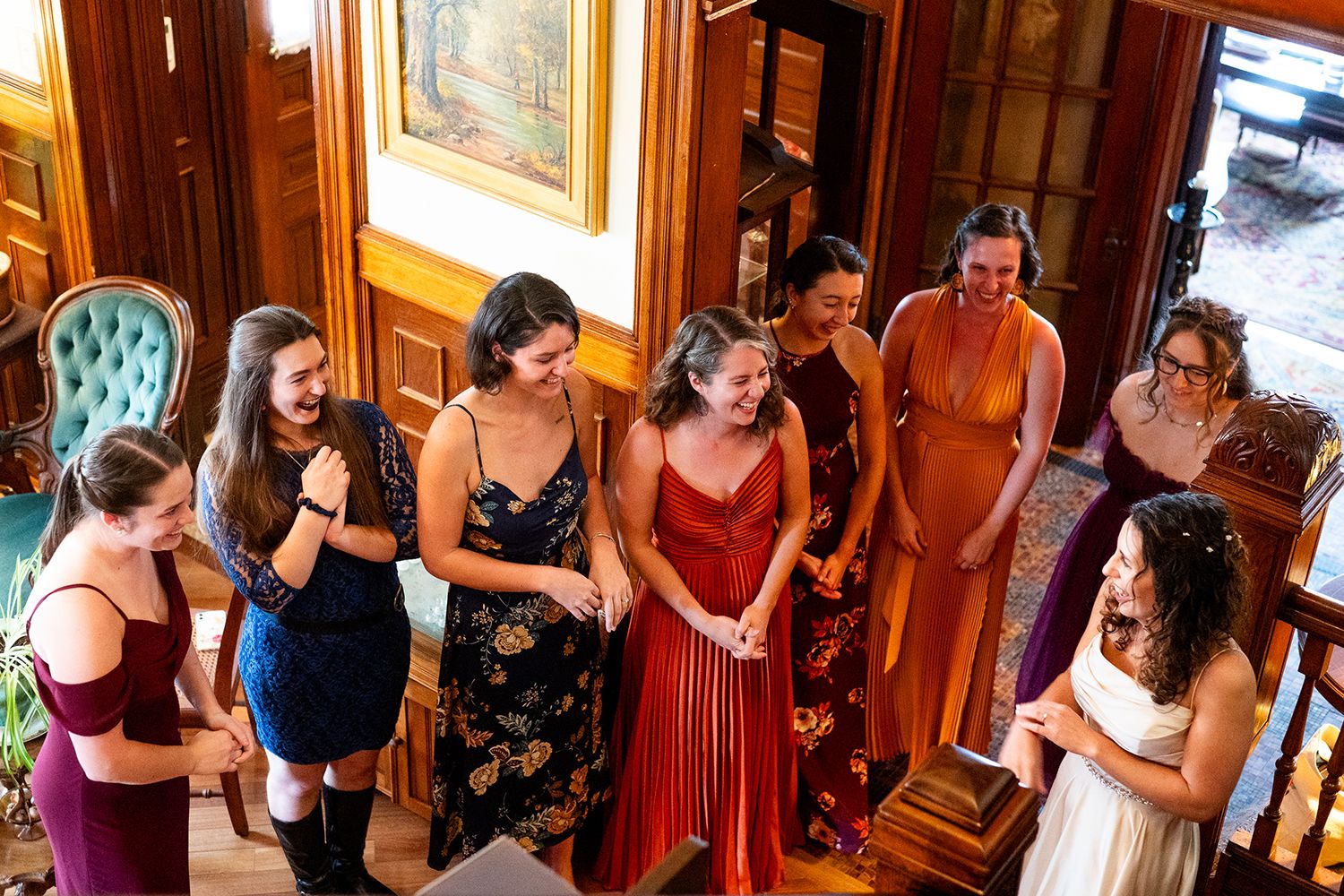 A group of wedding attendees in colorful dresses gather inside an elegant room with wooden walls and vintage decor.