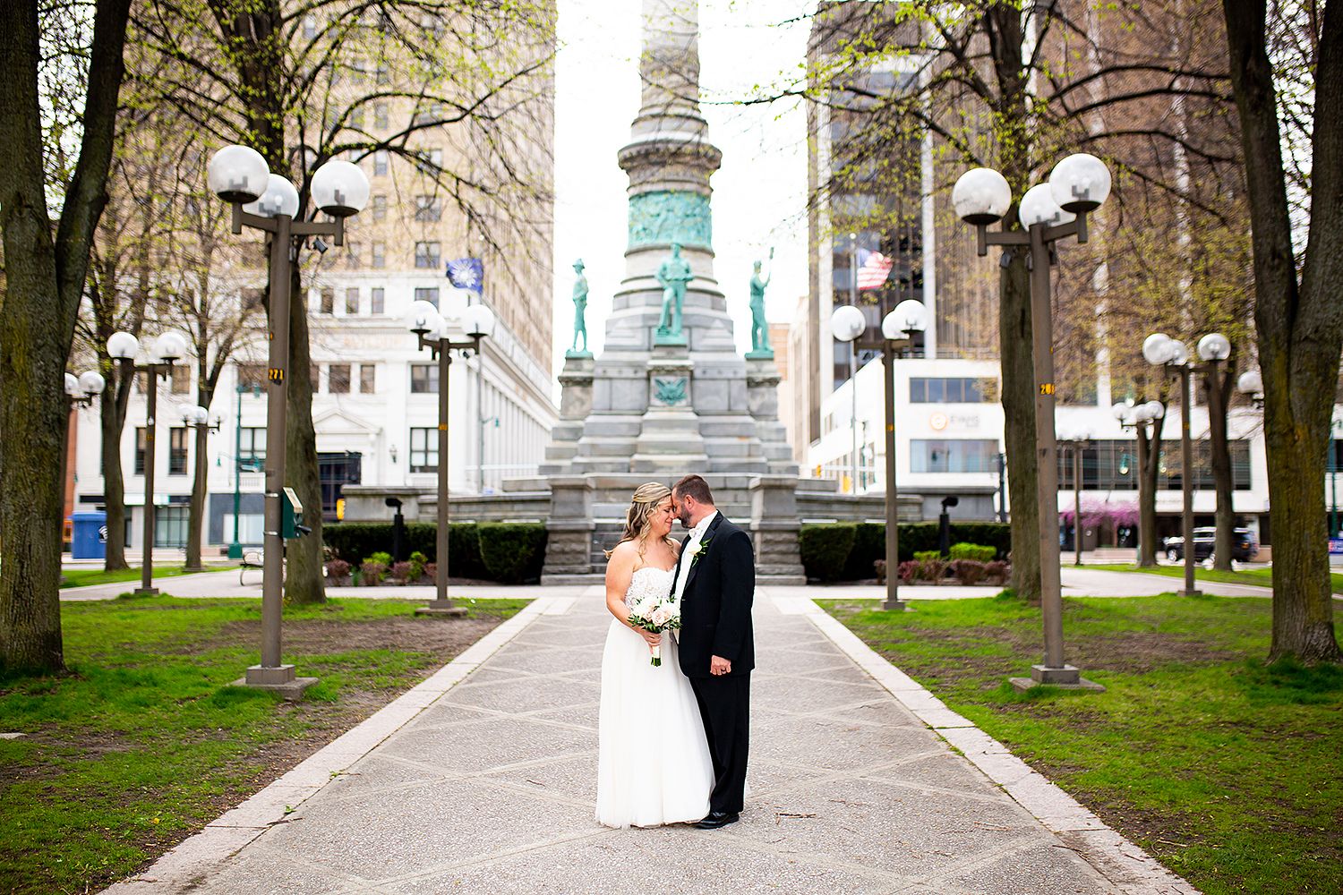 A couple shares a romantic wedding photoshoot in a tree-lined park with historic buildings in the background.