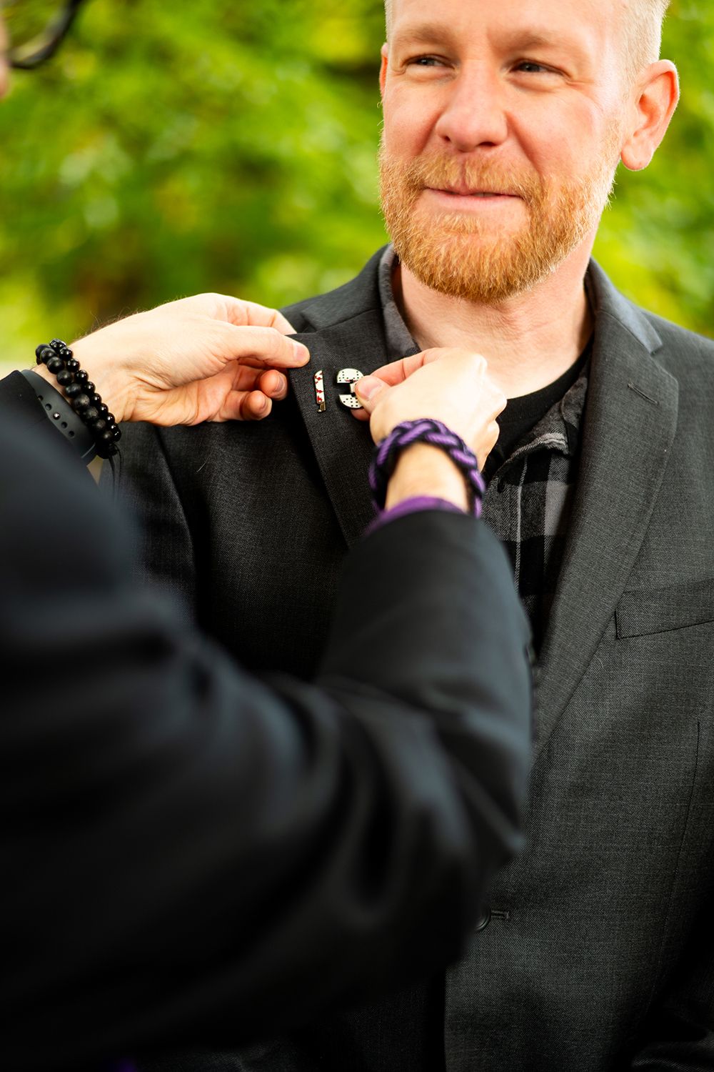 Someone adjusts a black suit lapel on a person wearing a dark blazer outdoors with green foliage in the background.