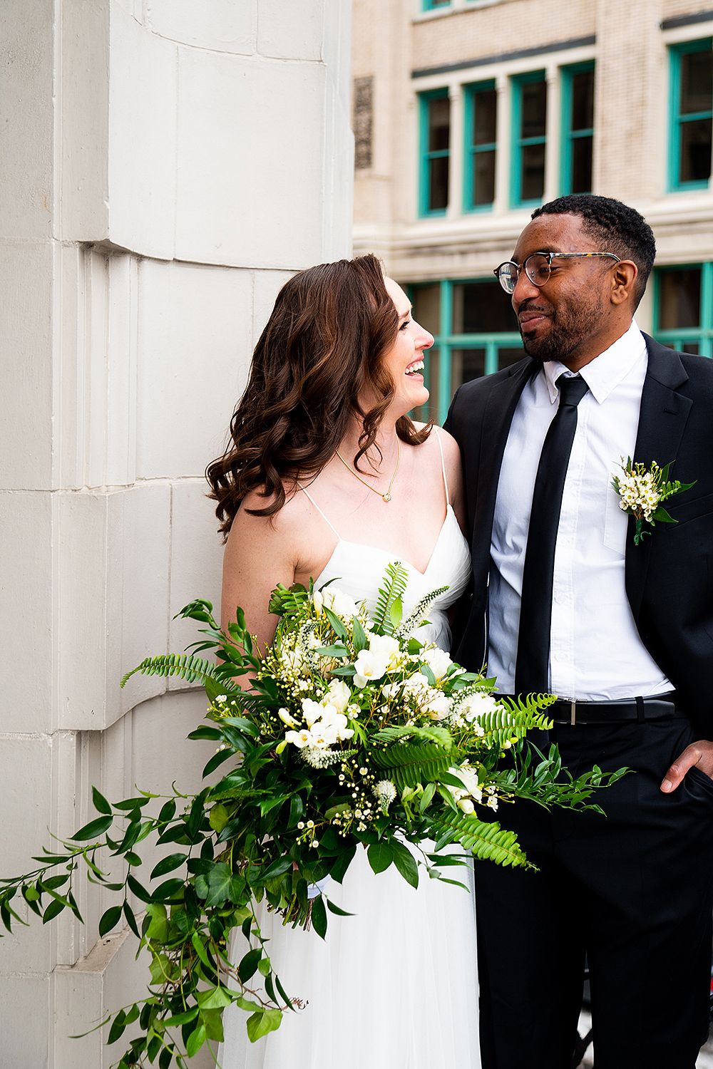 A wedding couple shares an intimate moment in a sequence of architectural columns with lush floral bouquet accents.