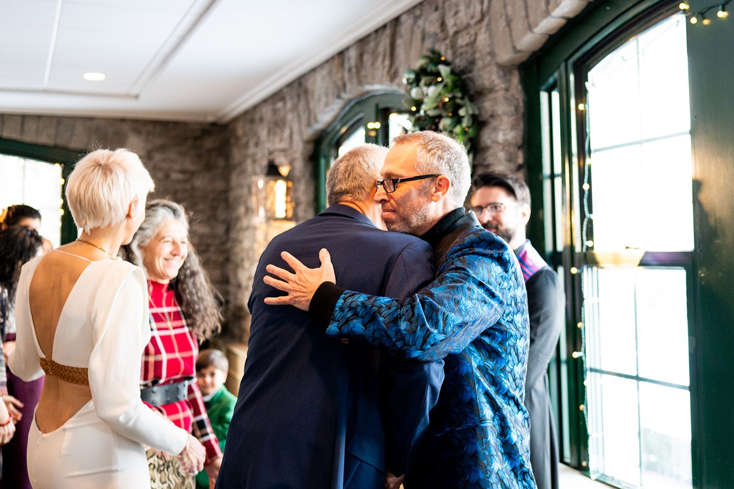 Two people share a warm embrace at an indoor celebration with exposed brick walls and guests in the background.