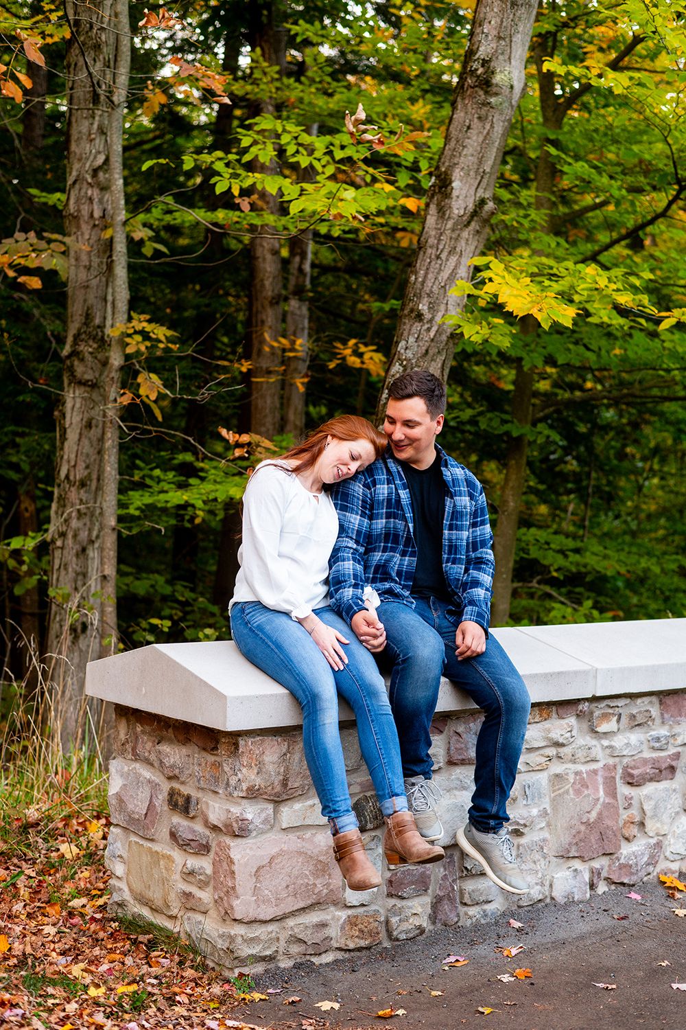 A couple sitting together on a stone wall in autumn, surrounded by colorful fall foliage in a forest setting.