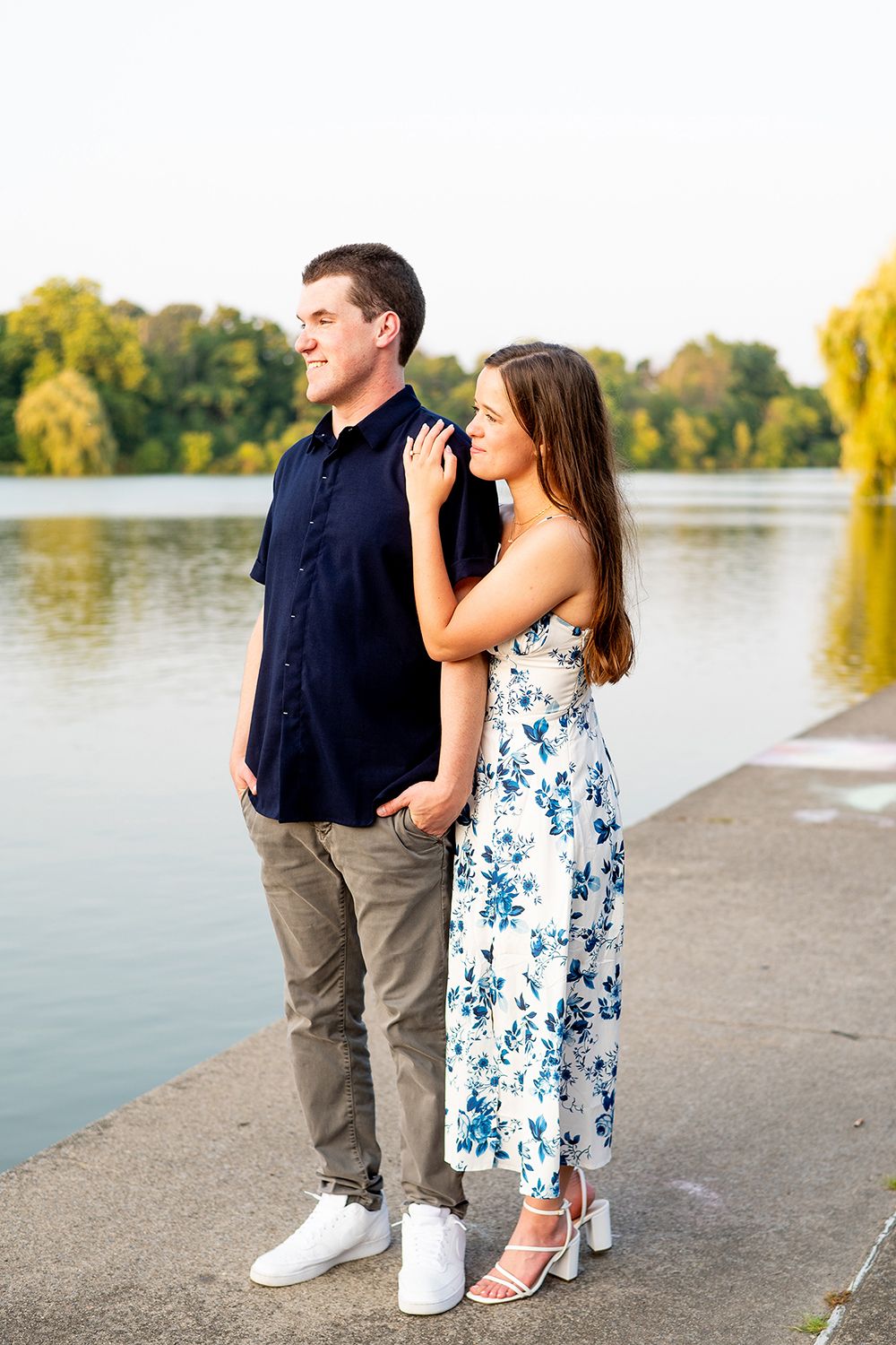A couple poses together by a lake at sunset, with her wearing a floral dress and him in a navy shirt.