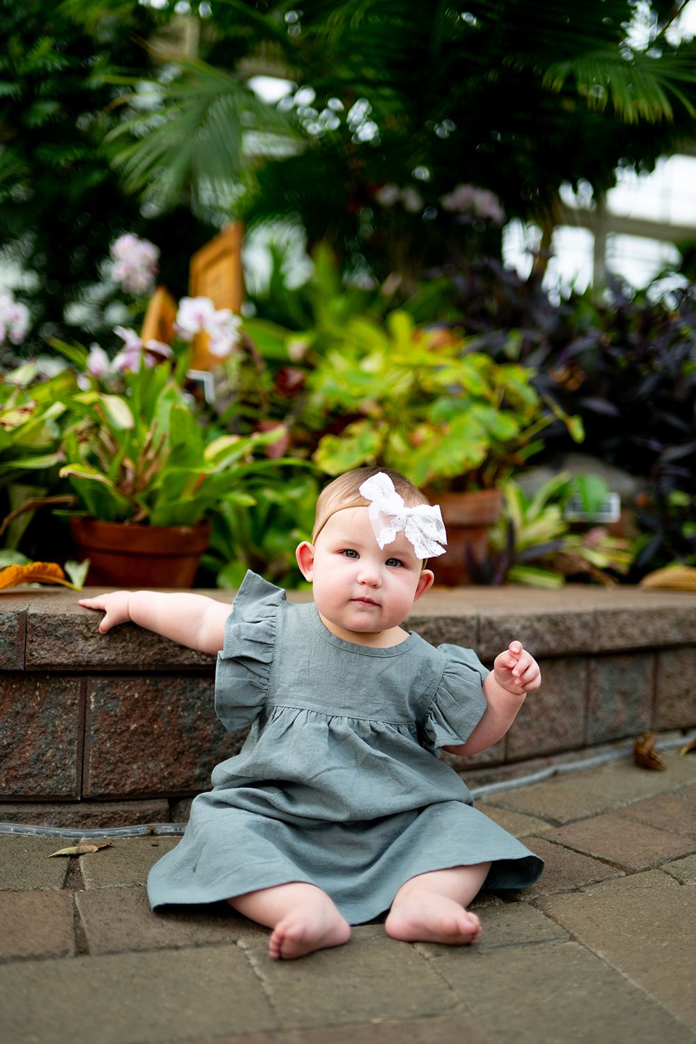 Small child in sage green dress sitting on stone patio surrounded by tropical plants.