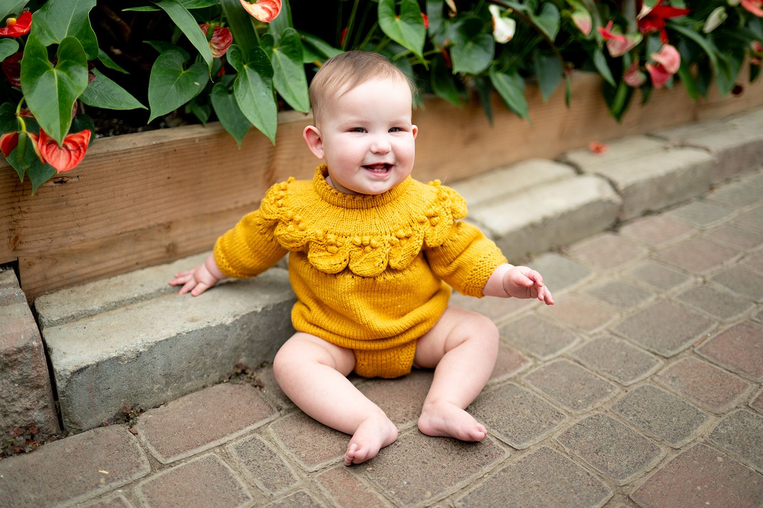 Baby wearing a mustard yellow ruffled top sits smiling on brick pavement near flowering red plants.